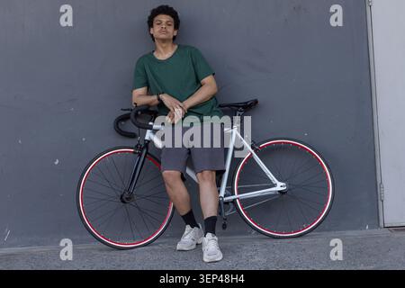 Homme africain adulte appuyé sur un vélo blanc noir à engrenage fixe sur le trottoir, portant un T-shirt vert foncé Banque D'Images