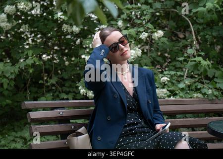 Jeune femme en lunettes de soleil se détend sur un banc en bois entouré de feuillage vert luxuriant et de fleurs blanches tout en tenant un smartphone. Communication numérique Banque D'Images