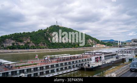 Les bateaux de croisière fluviale accostent sur le Danube à Budapest avec la colline Gellért et son monument de la liberté s'élevant au-dessus des pentes couvertes d'arbres. Drapeaux hongrois visib Banque D'Images
