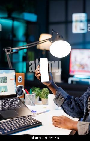 Individu afro-américain travaillant à la maison à un bureau avec ordinateur portable et smartphone avec écran blanc, jeune personne saisissant verticalement un appareil mobile affichant un modèle chromakey isolé. Banque D'Images
