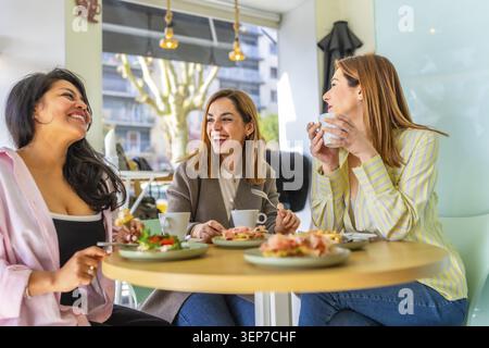 Trois amies diverses appréciant une réunion amusante et une conversation joyeuse autour d'un brunch sain et d'un café dans un cadre de café moderne et lumineux Banque D'Images