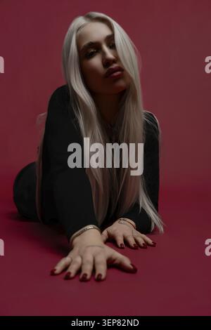 Une femme aux longs cheveux blonds repose gracieusement sur un fond rouge. Elle pose avec une expression détendue, ses mains pointant vers l'avant, améliorant l'arti Banque D'Images