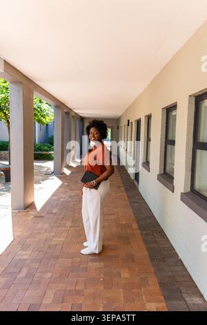 Femme afro-américaine debout dans le couloir portant un pantalon blanc blouse rouille tenant un pochette noire Banque D'Images