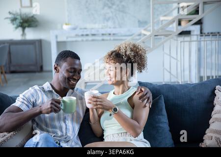 Couple afro-américain assis sur le canapé bleu penché ensemble dans la maison moderne tenant des tasses en céramique Banque D'Images