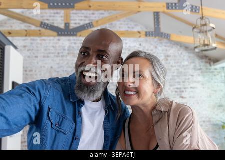 Couple souriant et posant pour selfie dans un loft rénové lumineux avec smartphone et suspension Banque D'Images