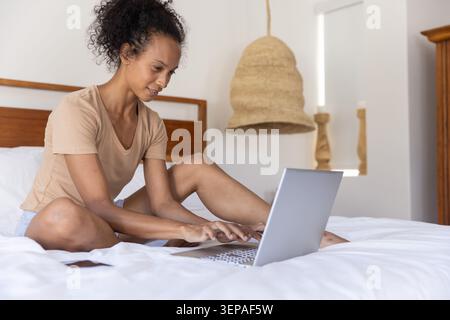 Femme afro-américaine et asiatique assise sur le lit tapant sur un ordinateur portable argenté à côté du smartphone à la maison Banque D'Images