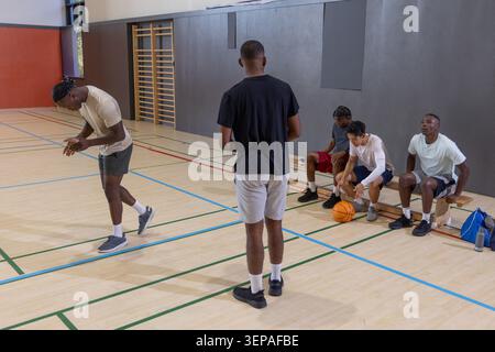 Divers coéquipiers masculins dans des chaussures de nouage d'équipement athlétique, assis sur un terrain en bois avec basket-ball Banque D'Images