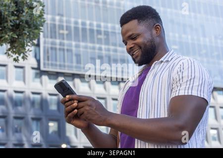 Homme afro-américain debout près d'un immeuble de bureaux, portant un t-shirt violet et tenant un smartphone Banque D'Images