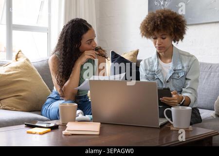 Amies afro-américaines examinant des papiers sur le canapé gris à l'aide d'un ordinateur portable et d'un téléphone Banque D'Images