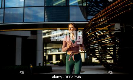 Une femme court sur un chemin près de grands bâtiments modernes dans un cadre urbain. Il est en fin d'après-midi, et le soleil projette des ombres sur le sol. La scène montre Banque D'Images