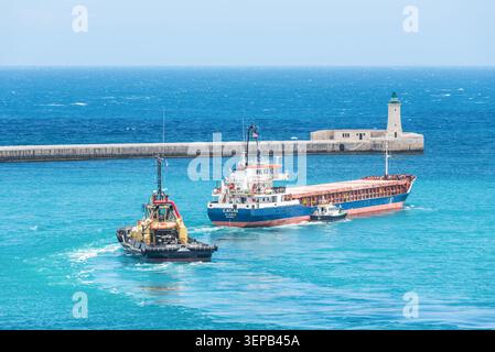 Valletta, Malte – 16 mai 2025 : cargo Ship, remorqueur et pilote naviguant près de l'entrée du port historique et phare de la Valette, Malte, surro Banque D'Images