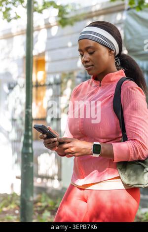 Femme africaine debout sur le trottoir urbain vérifiant le téléphone, portant montre et bandeau avec sac à dos Banque D'Images
