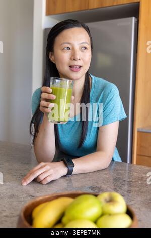 Femme asiatique mature penchée au comptoir de cuisine tenant un verre de smoothie vert avec un bol smartwatch Banque D'Images