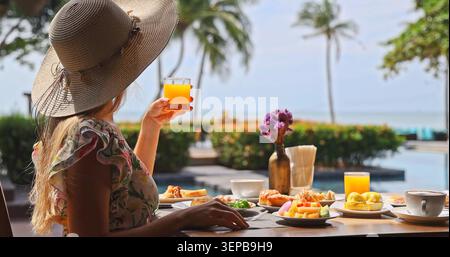Femme portant un chapeau de paille tenant un verre de jus d'orange à la table de petit déjeuner au bord de la piscine, vue sur l'océan tropical avec des palmiers, matin ensoleillé lumineux, trav de luxe Banque D'Images
