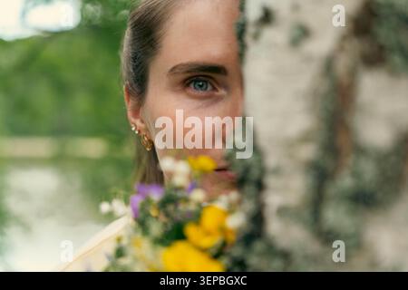 Gros plan portrait extérieur de femme avec un oeil visible, partiellement obscurci par un bouleau et un petit bouquet de fleurs colorées, avec naturel doux Banque D'Images