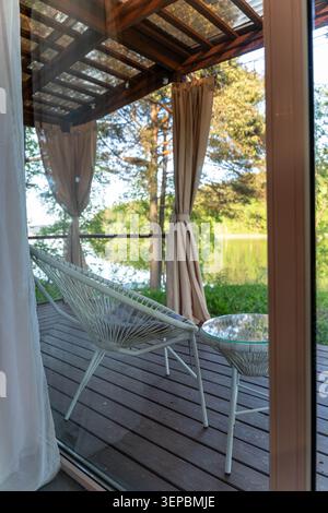 Terrasse couverte relaxante avec chaise longue tissée blanche, table ronde en verre, rideaux suspendus, terrasse en bois et vue calme sur le lac avec arbres et moelleux Banque D'Images