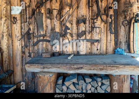 Ancien intérieur de hangar en bois avec établi altéré, bois de chauffage empilé, et collection d'outils à main suspendus sur le mur de planche rugueux, éclairé par la lumière du jour naturelle Banque D'Images