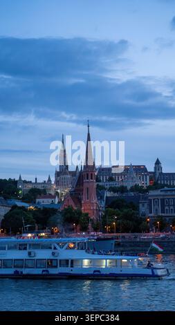 Horizon de Budapest au crépuscule avec l'église rouge emblématique et l'église Matthias le long du Danube avec bateau de tourisme au premier plan. réseau local hongrois Banque D'Images