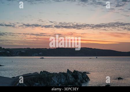 Ciel éclatant du matin aux couleurs pastel douces au-dessus de l'eau calme, avec un voilier ancré près d'une structure rocheuse sombre au premier plan à Looe Banque D'Images