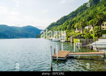 Quai en bois et jetée sur le rivage rocheux du lac de Lugano avec des montagnes vertes en arrière-plan, Caslano, canton du Tessin, Suisse. Jour d'été nuageux. NAT Banque D'Images