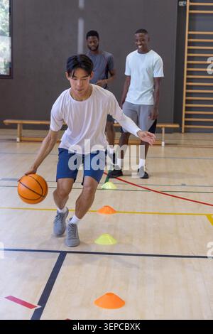 Divers coéquipiers masculins pratiquant l'exercice dribble dans la salle de gym, guidant le basket-ball orange à travers des cônes Banque D'Images