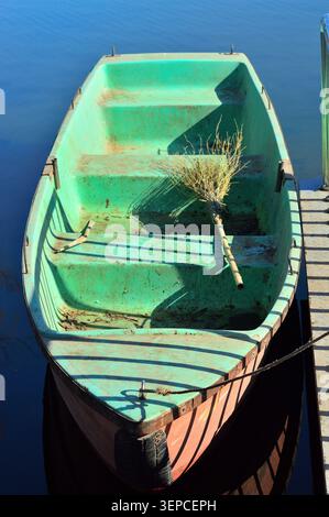 Vieille barque attachée à un quai en bois sur une eau bleue calme, vue d'en haut en plein soleil. Rustique et altérée, elle suggère un voyage tranquille au bord du lac. Banque D'Images