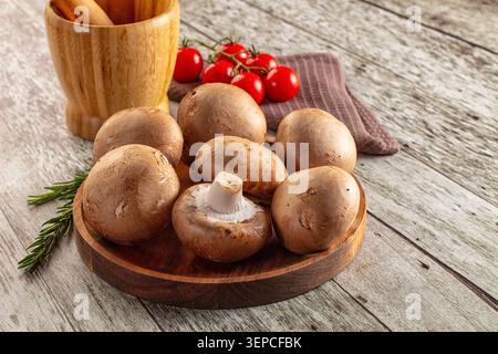 Champignons bruns frais disposés sur une planche à découper ronde en bois avec des tomates cerises sur la vigne un brin de romarin. Un pilon de mortier en bois se trouve dans th Banque D'Images