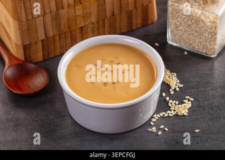 Gros plan de ramequin en céramique blanche rempli de pâte de tahini dorée lisse, de graines de sésame dispersées, d'une cuillère en bois et d'un pot en verre sur un comptoir en pierre sombre Banque D'Images