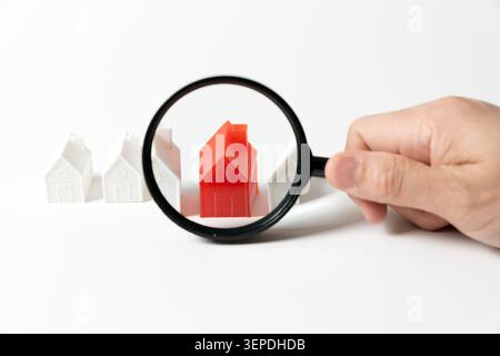 Une main tient une loupe au-dessus d'un modèle réduit de maison rouge unique dans une rangée de maisons en plastique blanc. Concept pour la recherche immobilière, l'inspection de propriété, la chasse à la maison Banque D'Images