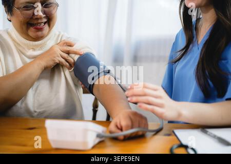 La femme aînée asiatique reçoit un contrôle de surveillance de la pression artérielle. Une jeune infirmière en gommage bleu ajuste l'appareil. Femme senior expression calme montre la confiance. Banque D'Images