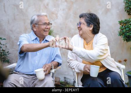 Couple de personnes âgées assis dans des chaises de jardin. Chacune contient une tasse de thé blanche. Ils sourient, joignant les mains en forme de cœur. Leur joie est claire et authentique. Idéal pour Banque D'Images