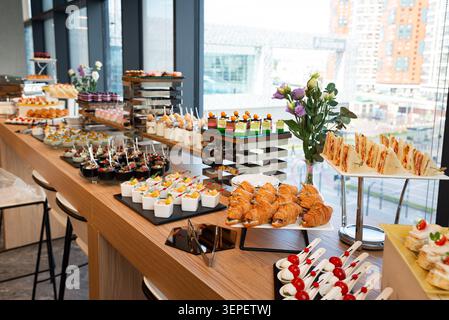 Variété de finger food et desserts colorés sur une table de buffet en bois dans un espace événementiel lumineux. Banque D'Images