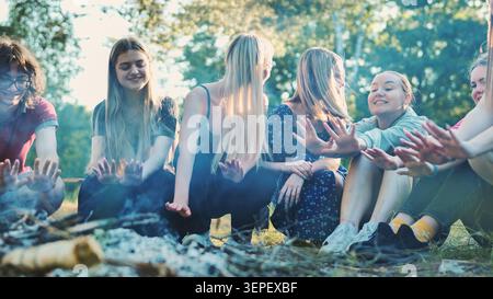 Groupe d'amis d'école heureux réchauffant les mains autour du feu de joie, profitant des vacances de camp d'été ensemble Banque D'Images