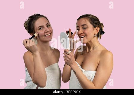 Deux jeunes femmes dans des serviettes appliquant le maquillage sur un fond rose Banque D'Images