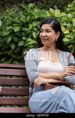 Femme assise sur un banc en bois tenant une tasse à emporter et un sac en cuir marron reposant dans le parc Banque D'Images