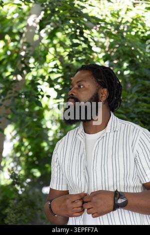 Homme africain adulte ajustant chemise rayée tout en se tenant dans le jardin portant T-shirt blanc, bracelet Banque D'Images