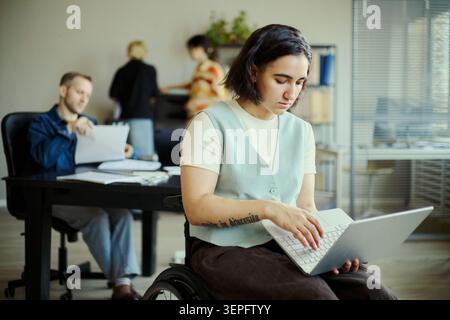 Jeune femme adulte handicapée utilisant un ordinateur portable au bureau tandis que des collègues collaborent en arrière-plan, représentant un lieu de travail inclusif. Idéal pour les campagnes RH, DEI, embauche, travail d'équipe Banque D'Images