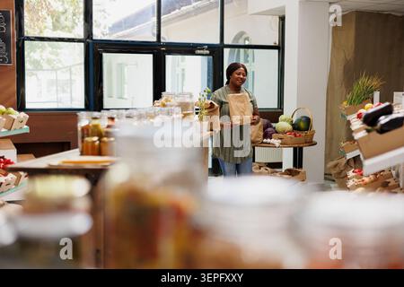 Femme noire recherche des produits cultivés localement dans un supermarché, parcourant des produits écologiques en mettant l'accent sur le fond ; client marchant dans un magasin de bio-aliments tout en transportant un sac en papier brun. Banque D'Images