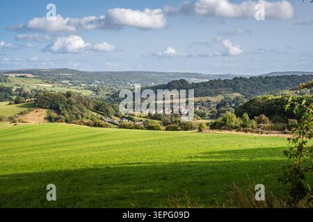 Large paysage horizontal vue sur les champs verdoyants ensoleillés vers le village historique de Stoney Middleton niché dans la vallée boisée de th Banque D'Images