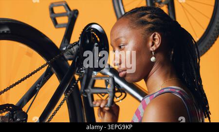 Femme afro-américaine souriante satisfaite tout en poussant les pédales pour faire tourner la roue de vélo, après avoir réussi à la réparer, professionnel satisfait des résultats après avoir ajusté le bras de manivelle de vélo défectueux. Banque D'Images