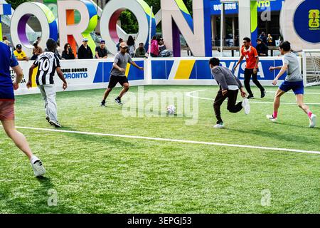 Toronto, Canada - le 14 juin 2026 : un groupe de jeunes hommes divers, y compris des joueurs asiatiques, noirs et blancs, participent à un tournoi interactif de mini-football au Nathan Phillips Square. Capturés en pleine action dribbles et en passant un ballon de soccer sur un terrain escamotable, ils jouent sur le fond emblématique de Toronto. Entourée d'une image de marque dynamique, cette scène animée met en valeur l'engagement des fans et l'esprit communautaire lors des célébrations officielles de la Coupe du monde de la FIFA 2026 à Toronto. Banque D'Images