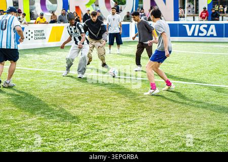 Toronto, Canada - le 14 juin 2026 : un groupe de jeunes hommes divers, y compris des joueurs asiatiques, noirs et blancs, participent à un tournoi interactif de mini-football au Nathan Phillips Square. Capturés en pleine action dribbles et en passant un ballon de soccer sur un terrain escamotable, ils jouent contre l'emblématique hôtel de ville de Toronto en toile de fond. Entourée d'une image de marque dynamique, cette scène animée met en valeur l'engagement intense des fans et l'esprit communautaire lors des célébrations officielles de la Coupe du monde de la FIFA 2026 à Toronto. Banque D'Images