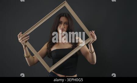 Femme tenant un cadre vide autour de son visage contre un fond de studio sombre Banque D'Images