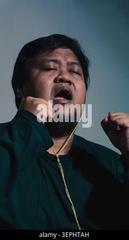 Un homme adulte indonésien expressif portant une chemise vert foncé chante passionnément les yeux fermés et les poings serrés tout en portant des écouteurs filaires contre Banque D'Images