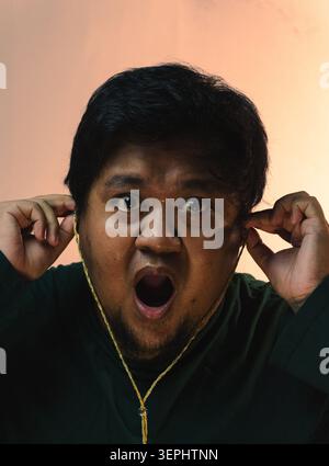 Un homme indonésien portant une chemise vert foncé insère des écouteurs filaires dans ses oreilles tout en regardant l'appareil photo avec une expression intense choquée contre A. Banque D'Images