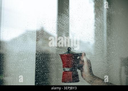 Main tenant un pot de moka rouge derrière un verre texturé humide avec des gouttelettes d'eau. Scène d'accueil atmosphérique sur le rituel du café du matin, style de vie confortable, Li lent Banque D'Images