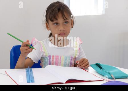 Fille assise au bureau de l'école dans une robe rayée, tenant un crayon vert, un cahier, une règle bleue Banque D'Images
