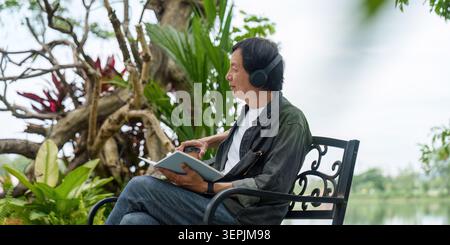 Homme asiatique senior portant des écouteurs lisant le livre dans le parc à l'extérieur pour la détente et le vieillissement actif Banque D'Images