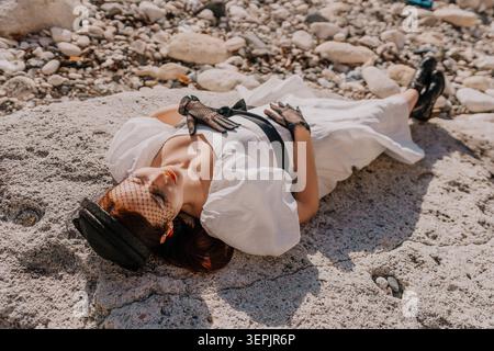 Femme robe côte élégante dame portant une robe blanche et voile noir couché sur des pierres côtières en plein soleil Banque D'Images