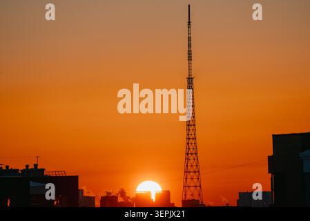 Silhouette de tour de radio s'élevant au-dessus des bâtiments de la ville au coucher du soleil, avec un ciel orange et jaune vibrant formant une toile de fond colorée, représentant la vie urbaine moderne et la connectivité technologique Banque D'Images